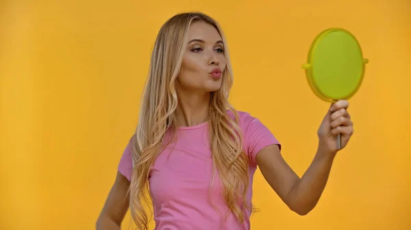 Mulher loira amamentando lábios enquanto segurando espelho isolado no amarelo — Fotografia de Stock