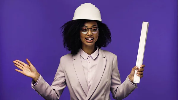 Cheerful african american engineer with braces holding rolled paper isolated on purple — Stock Photo