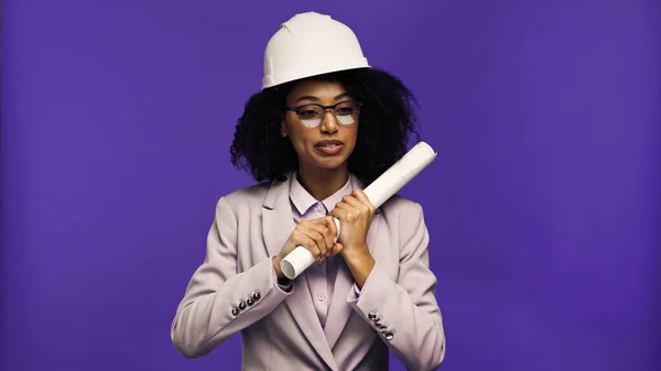 African american engineer in safety helmet holding rolled paper isolated on purple — Stock Photo