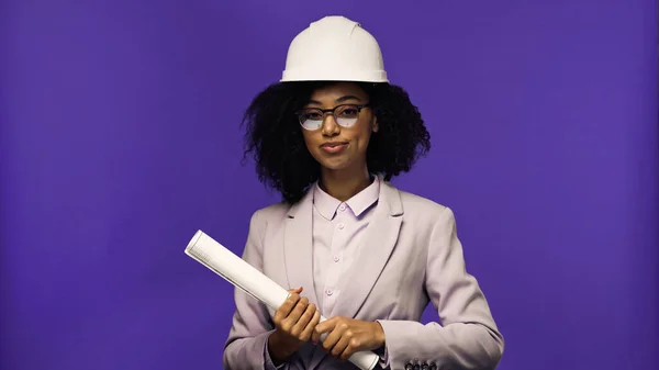 Complacido ingeniero afroamericano en gafas y casco de seguridad sosteniendo papel laminado aislado en púrpura - foto de stock