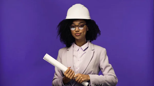 Engenheiro americano africano satisfeito em óculos e capacete de segurança segurando papel laminado isolado em roxo — Fotografia de Stock