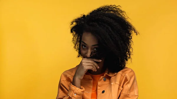 Junge und schüchterne afrikanisch-amerikanische Frau bedeckt Gesicht mit lockigem Haar isoliert auf gelb — Stockfoto