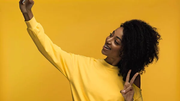 Весела афроамериканка з брекетами, що показують знак миру, приймаючи селфі ізольовано на жовтому — стокове фото