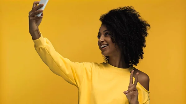 Mujer afroamericana feliz con frenos que muestran signo de paz mientras toma selfie aislado en amarillo - foto de stock
