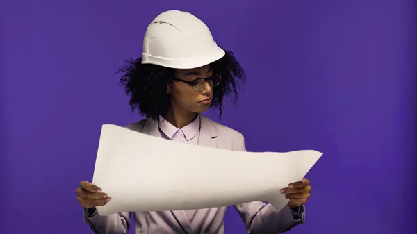 Mujer afroamericana rizada en gafas y casco de seguridad mirando el plano aislado en púrpura - foto de stock