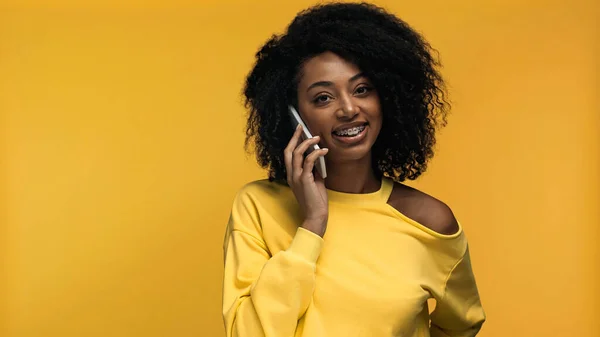 Allegra donna afroamericana con bretelle che parla su smartphone isolato su giallo — Foto stock