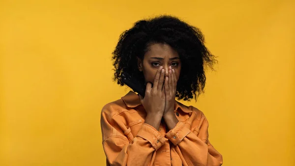 Triste africano americano mujer cubierta boca mirando cámara aislado en amarillo - foto de stock