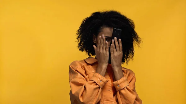 Напуганная африканская американка покрывает лицо и держит пульт дистанционного управления изолирован на желтый — стоковое фото