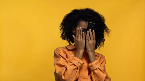 Mulher afro-americana assustada cobrindo rosto e segurando controlador remoto isolado no amarelo — Fotografia de Stock