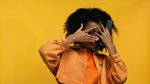 Scared african american woman with braces covering face and holding remote controller isolated on yellow — Stock Photo
