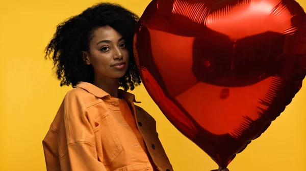 Femme afro-américaine frisée près ballon rouge en forme de coeur isolé sur jaune — Photo de stock