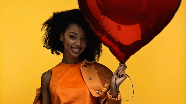 Веселая африканская американка с брекетами, держащими красный шар в форме сердца, изолированный на желтом — стоковое фото