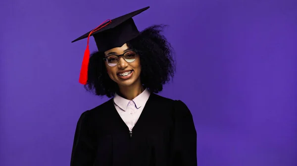 Cheerful african american student with braces in graduation cap and gown isolated on purple — Stock Photo