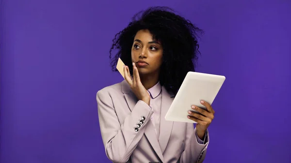 Joven afroamericana mujer en chaqueta sosteniendo tableta digital y tarjeta de crédito aislado en púrpura - foto de stock