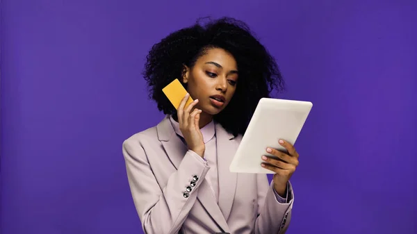 Joven afroamericana mujer en chaqueta usando tableta digital y la celebración de tarjeta de crédito aislado en púrpura - foto de stock