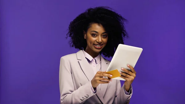 Alegre afroamericana mujer en chaqueta celebración digital tableta y tarjeta de crédito aislado en púrpura - foto de stock