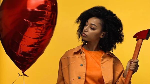 Curly african american woman holding hammer near red heart-shaped balloon isolated on yellow, valentines day concept — Stock Photo