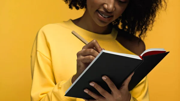 Обрізаний вид щасливої афроамериканської жінки з брекетами, що пишуть в блокноті ізольовані на жовтому — стокове фото