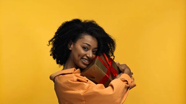 Alegre afroamericana mujer con brackets abrazo envuelto presente aislado en amarillo - foto de stock