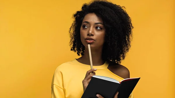 Mujer afroamericana pensativa sosteniendo pluma y cuaderno aislado en amarillo - foto de stock