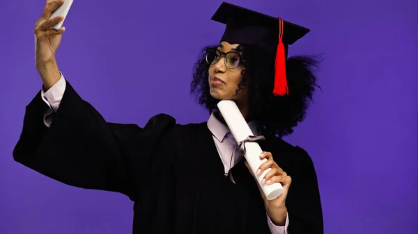 Joven africano americano estudiante en gafas y graduación gorra tomando selfie aislado en púrpura - foto de stock