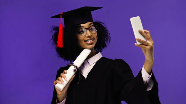 Cheerful african american student with braces in graduation cap and gown taking selfie with rolled diploma isolated on purple — Stock Photo