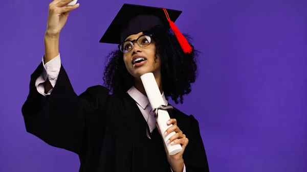 Happy african american student with braces in graduation cap and gown taking selfie with rolled diploma isolated on purple — Stock Photo