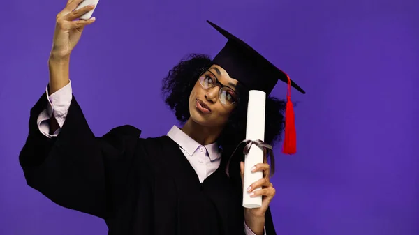 Smiling african american student in graduation cap and gown taking selfie with rolled diploma isolated on purple — Stock Photo