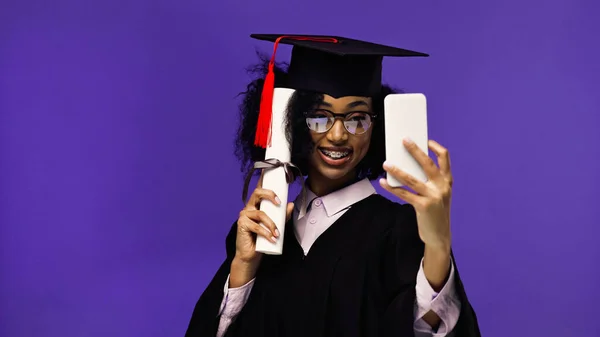 Smiling african american student with braces in graduation cap and gown taking selfie with diploma isolated on purple — Stock Photo