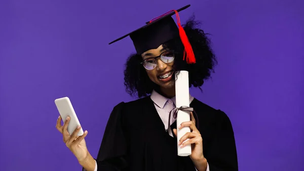 Estudiante afroamericano sonriente con frenos en gorra de graduación y vestido con teléfono inteligente y diploma aislado en púrpura - foto de stock