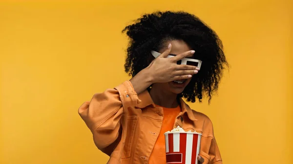 Verängstigte junge afrikanisch-amerikanische Frau in Hosenträgern und 3D-Gläsern mit Popcorn-Eimer isoliert auf gelb — Stockfoto
