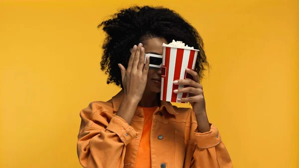 Напуганная африканская американка в 3D очках, держащая ведро попкорна и смотрящая фильм, изолированный на желтом — стоковое фото