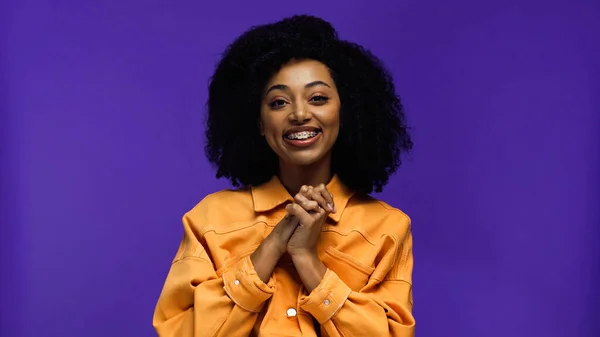 Femme afro-américaine gaie avec bretelles debout avec les mains serrées isolées sur violet — Photo de stock