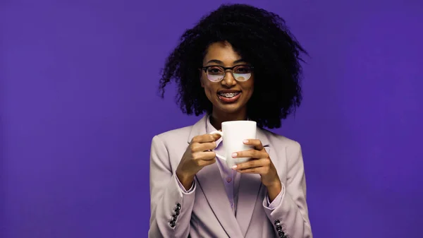 Cheerful african american businesswoman with braces holding cup of coffee isolated on purple — Stock Photo