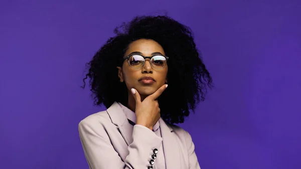 Curly african american businesswoman in glasses and blazer looking at camera isolated on purple — Stock Photo