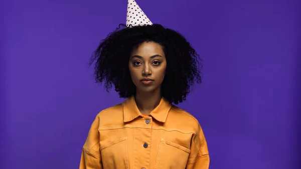 Young african american woman in party cap looking at camera isolated on purple — Stock Photo