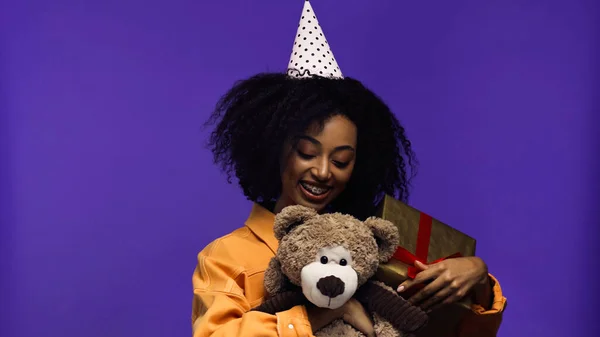 Fröhliche afrikanisch-amerikanische Frau mit Hosenträgern und Party-Mütze mit verpacktem Geschenkkarton und Teddybär isoliert auf lila — Stockfoto