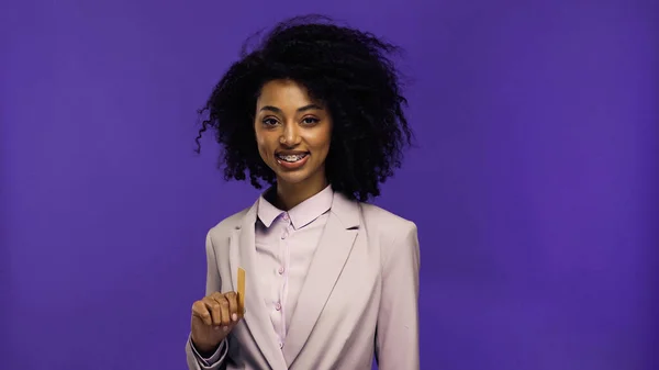 Cheerful african american businesswoman with braces holding credit card isolated on purple — Stock Photo