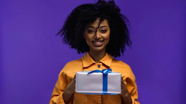 Щаслива афроамериканська жінка з брекетами, що тримає загорнутий подарунок ізольовано на фіолетовому — стокове фото