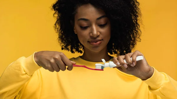 African american woman squeezing toothpaste on toothbrush isolated on yellow — Stock Photo