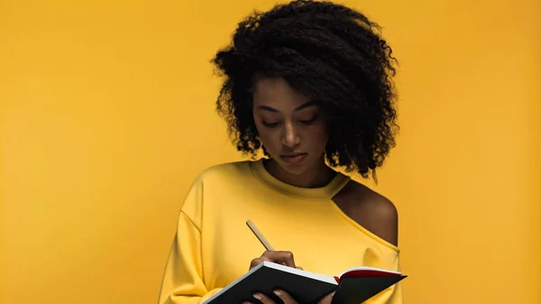 Кучерява афроамериканська жінка, що пише в блокноті ізольована на жовтому — стокове фото