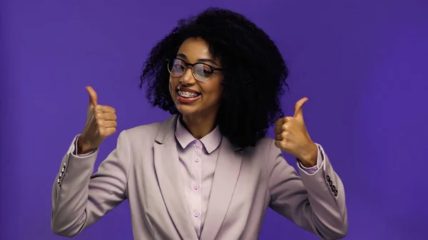 Feliz afroamericana mujer de negocios en gafas y chaqueta mostrando pulgares hacia arriba aislado en púrpura - foto de stock