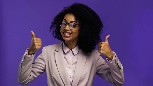 Felice donna d'affari africana americana in occhiali e blazer mostrando pollici in alto isolato su viola — Foto stock