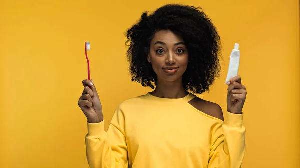 Усміхнена афроамериканка тримає зубну пасту з зубною щіткою ізольовано на жовтому — стокове фото