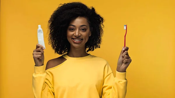 Весела афроамериканка з брекетами, що тримають зубну пасту з зубною щіткою ізольовано на жовтому — стокове фото