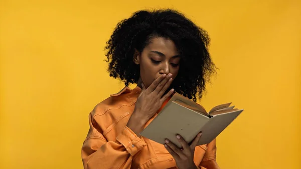Afro-américaine femme couvrant la bouche tout en lisant livre isolé sur jaune — Photo de stock