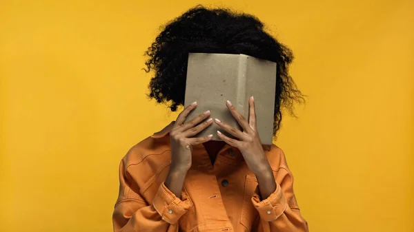 Mujer afroamericana cubriendo la cara con libro aislado en amarillo - foto de stock