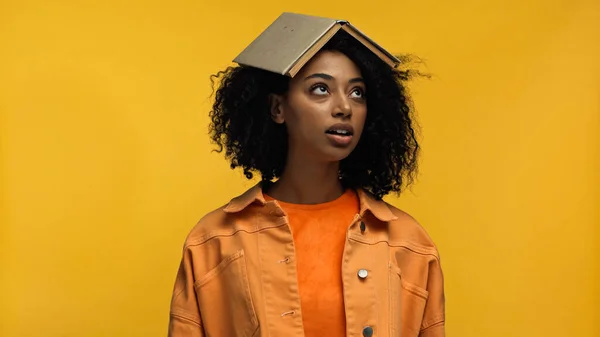 Jeune femme afro-américaine avec bretelles et livre sur la tête regardant isolé sur jaune — Photo de stock