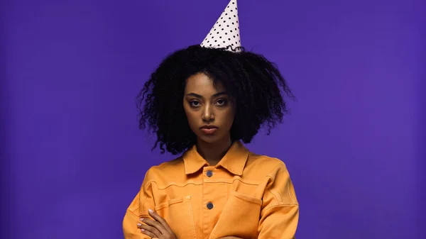 Mujer afroamericana seria en gorra de fiesta de pie con los brazos cruzados y mirando a la cámara aislada en púrpura - foto de stock