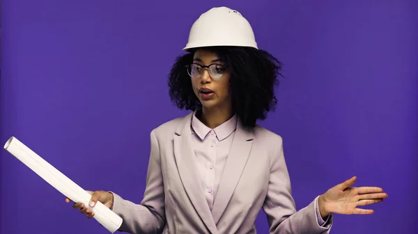 Mujer afroamericana disgustada en casco de seguridad sosteniendo papel laminado aislado en púrpura - foto de stock